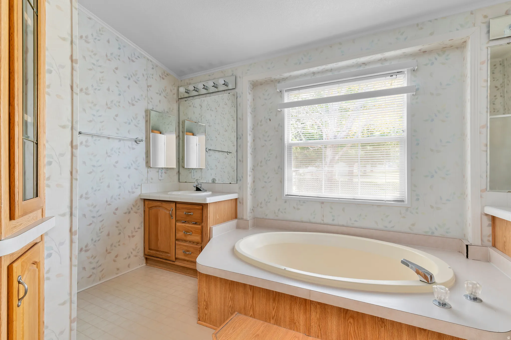 Spa-inspired primary bathroom featuring a sunken oval soaking tub with integrated deck jets, wood-finish cabinetry vanity with dual sinks, marble-look tile walls, polished nickel fixtures, and a large picture window flooding the space with natural light