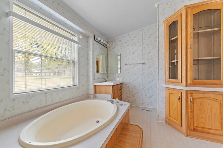 Spacious bath featuring an oval soaking tub, a double window with blinds, a vanity with a mirror and strip lighting, and a built-in wood-finish cabinet with glass inserts