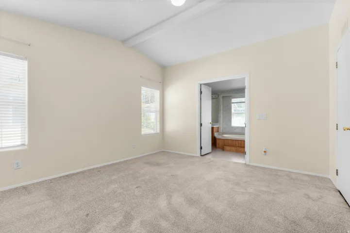 Spacious room featuring vaulted ceilings, two windows with blinds, light-colored carpeting, and a doorway opening to a bathroom with a built-in tub