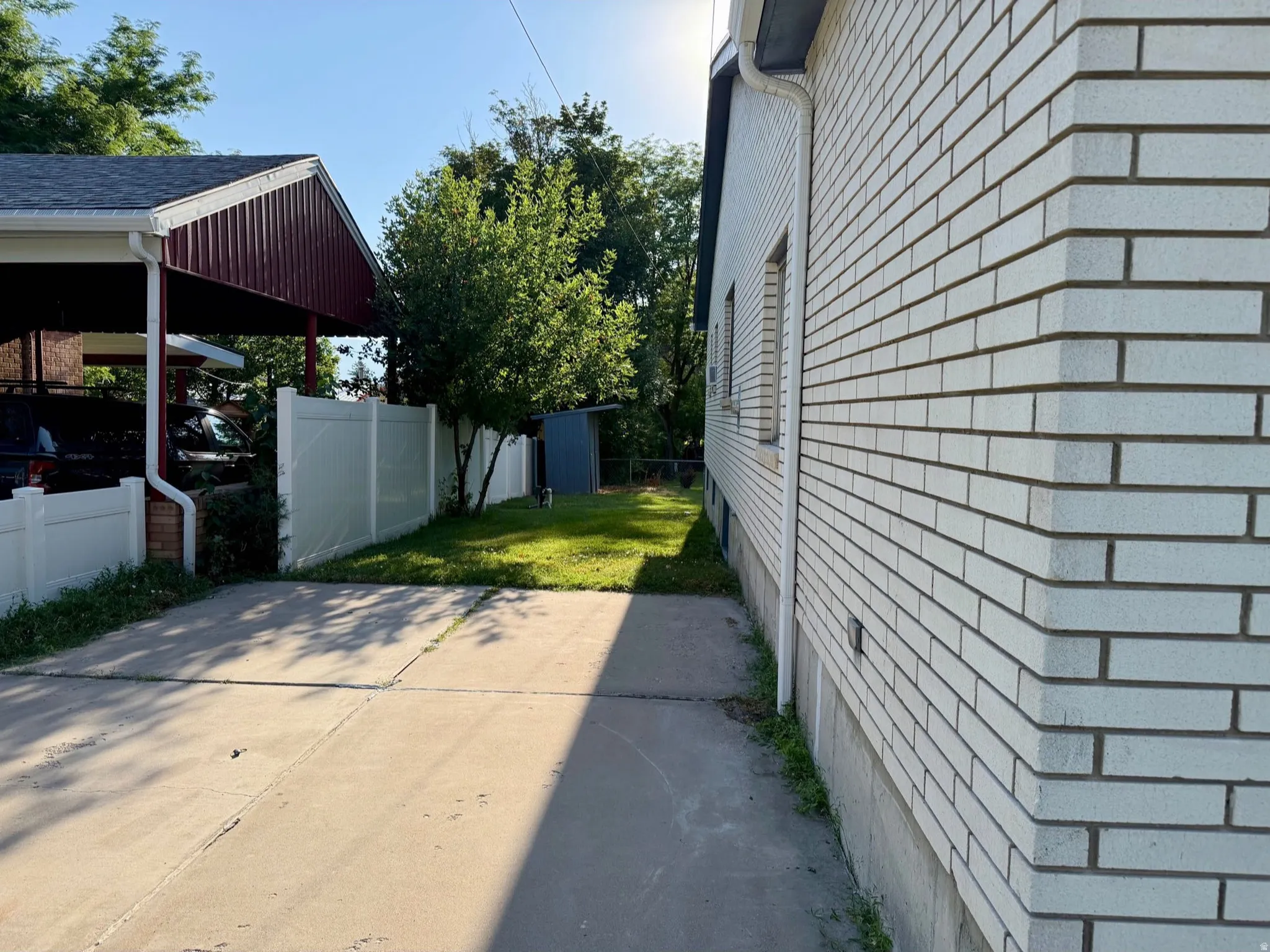 White brick exterior facade and siding, concrete driveway, lush green lawn, mature trees, and privacy fencing