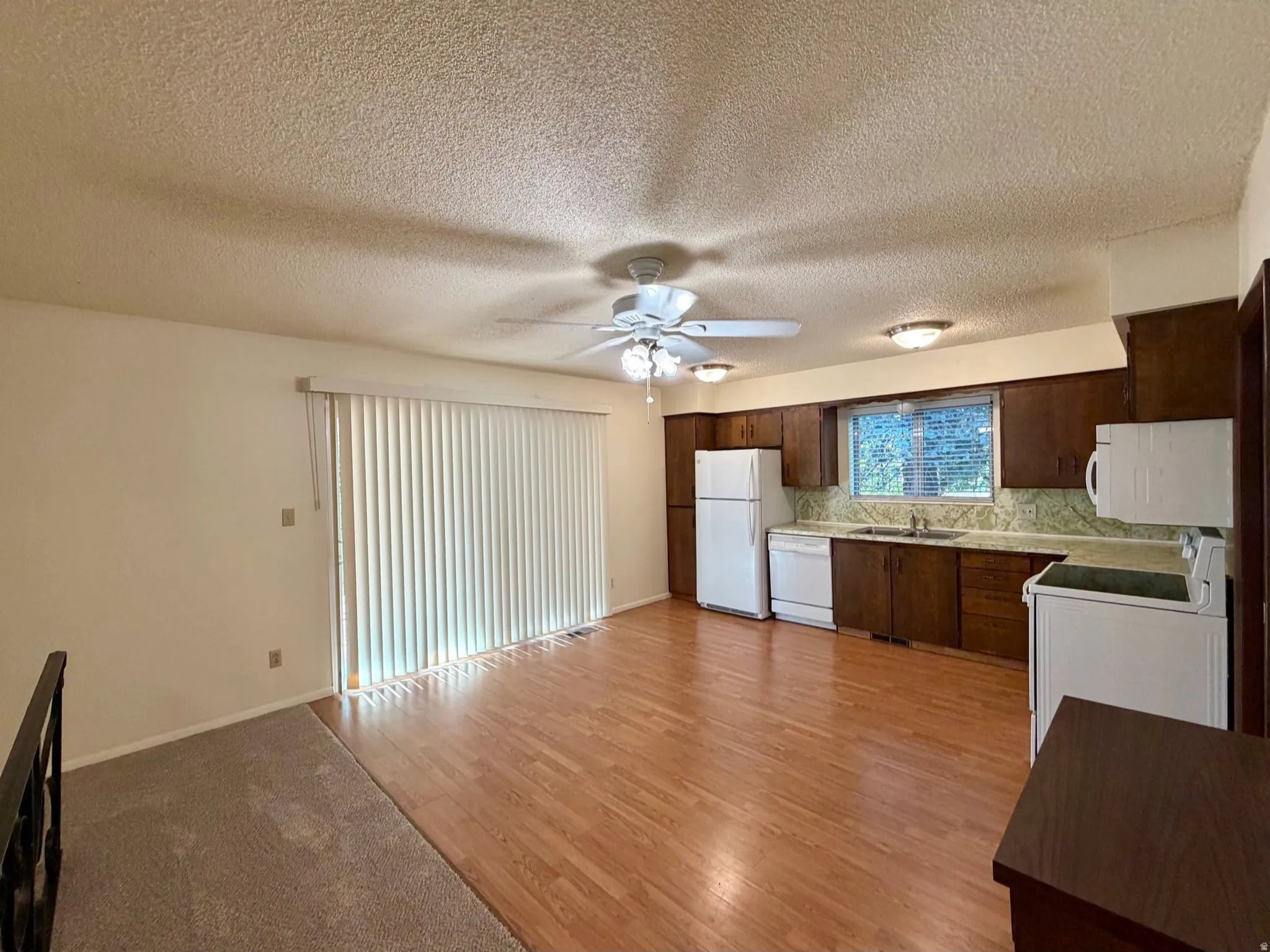 Kitchen and living area featuring wood-finish flooring, a ceiling fan with integrated lighting, a window with horizontal blinds, a kitchen with white appliances, and wood cabinetry
