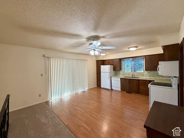 Kitchen and living area featuring wood-finish flooring, a ceiling fan with integrated lighting, a window with horizontal blinds, a kitchen with white appliances, and wood cabinetry