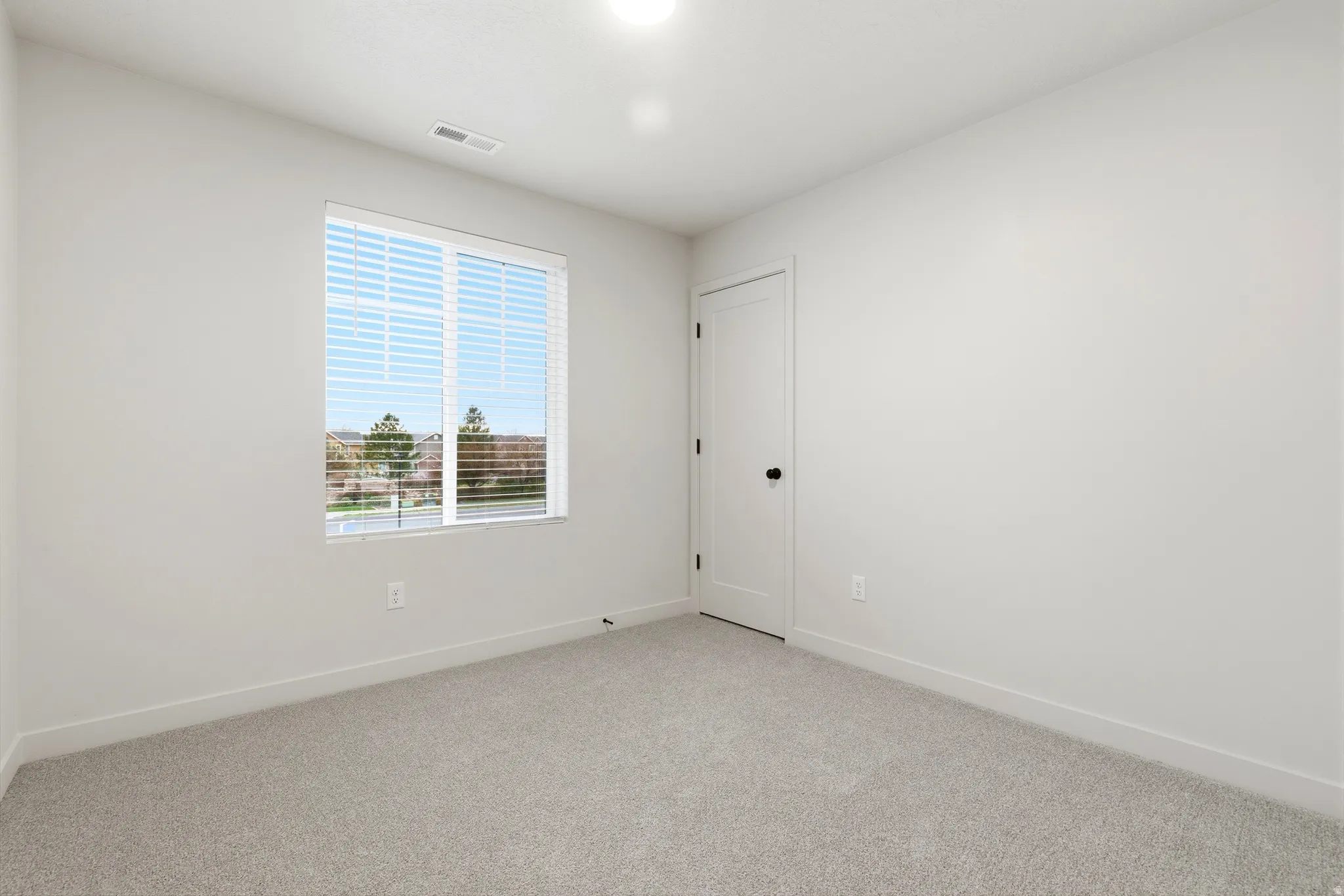 Neutral-toned room featuring a single window with blinds, light gray wall-to-wall carpeting, white walls, and a white interior door with black hardware