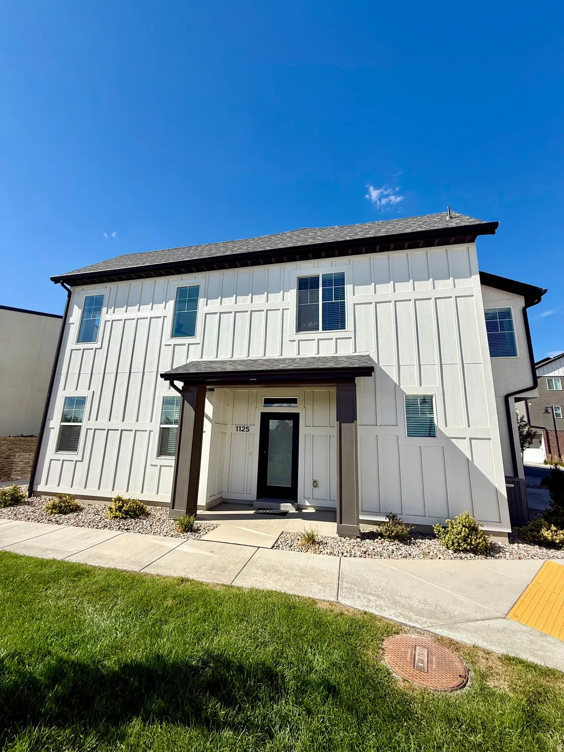 White board and batten siding, dark-framed windows, covered entry porch with dark columns, dark asphalt shingle roof, and a concrete walkway