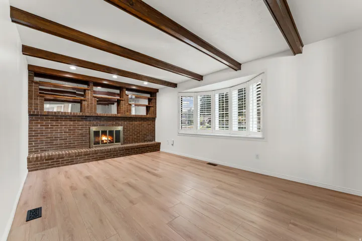 Spacious living room featuring exposed wooden ceiling beams, wide-plank wood-finish flooring, and a brick fireplace with built-in shelving and cabinetry