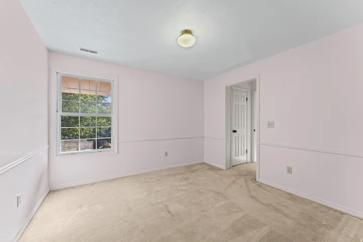 Bright and spacious bedroom featuring soft pink walls with crisp white chair rail molding, carpeted flooring, large double-hung window with natural light, ceiling-mounted light fixture, and direct access to an en-suite bathroom with white six-panel door