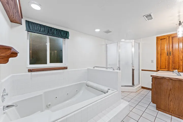 Spa-inspired primary bathroom featuring a deep soaking tub with whirlpool jets, separate walk-in shower enclosure with glass doors, dual vanity with wood cabinetry, ceramic tile flooring, recessed lighting, and a window with custom valance