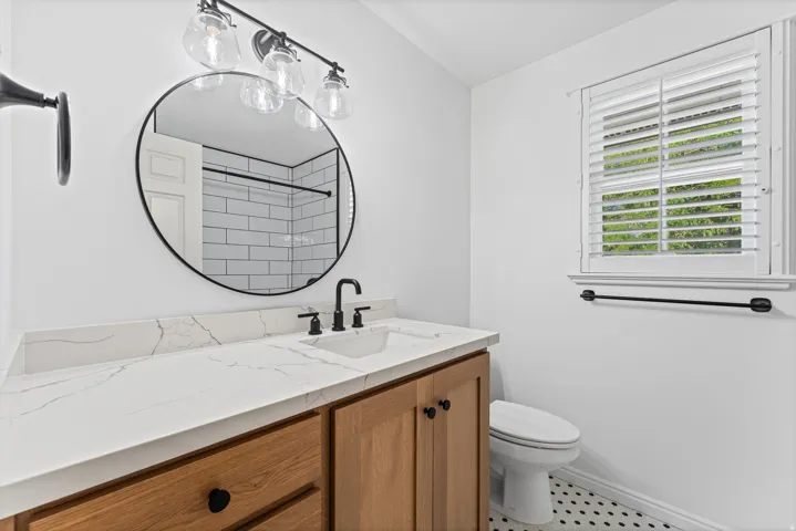 Spa-inspired bathroom featuring a circular black-framed mirror, white subway tile backsplash, wood-finish vanity with ample storage, white quartz countertop, matte black faucet and hardware, and white plantation shutters framing a bright window