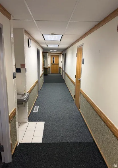 Well-maintained commercial corridor featuring polished drop ceiling with recessed lighting, dual wood-trimmed doorways with natural finish frames, and durable low-pile carpeting with contrasting black entry mat