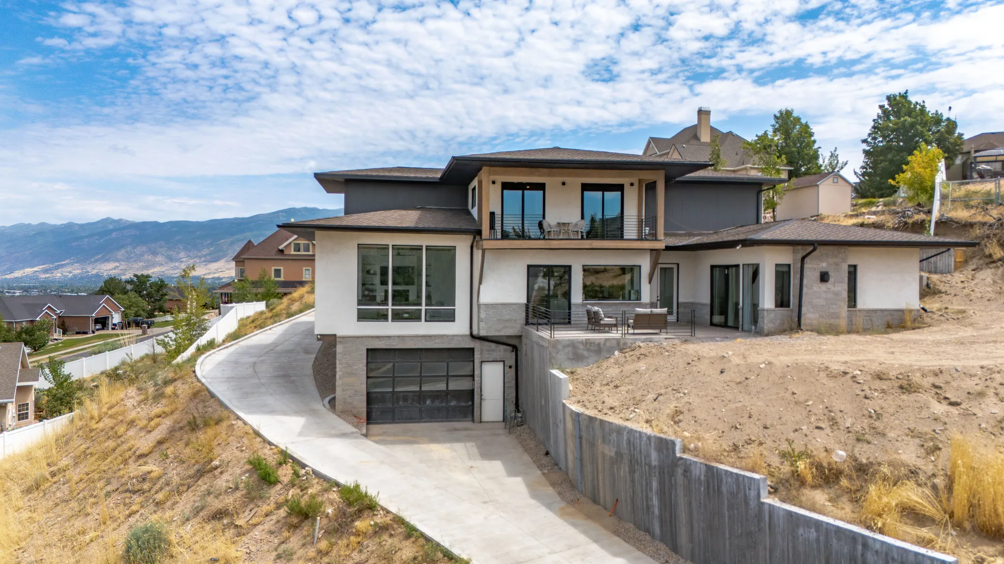 Modern architectural exterior featuring multiple balconies, expansive windows, a concrete driveway, and a retaining wall