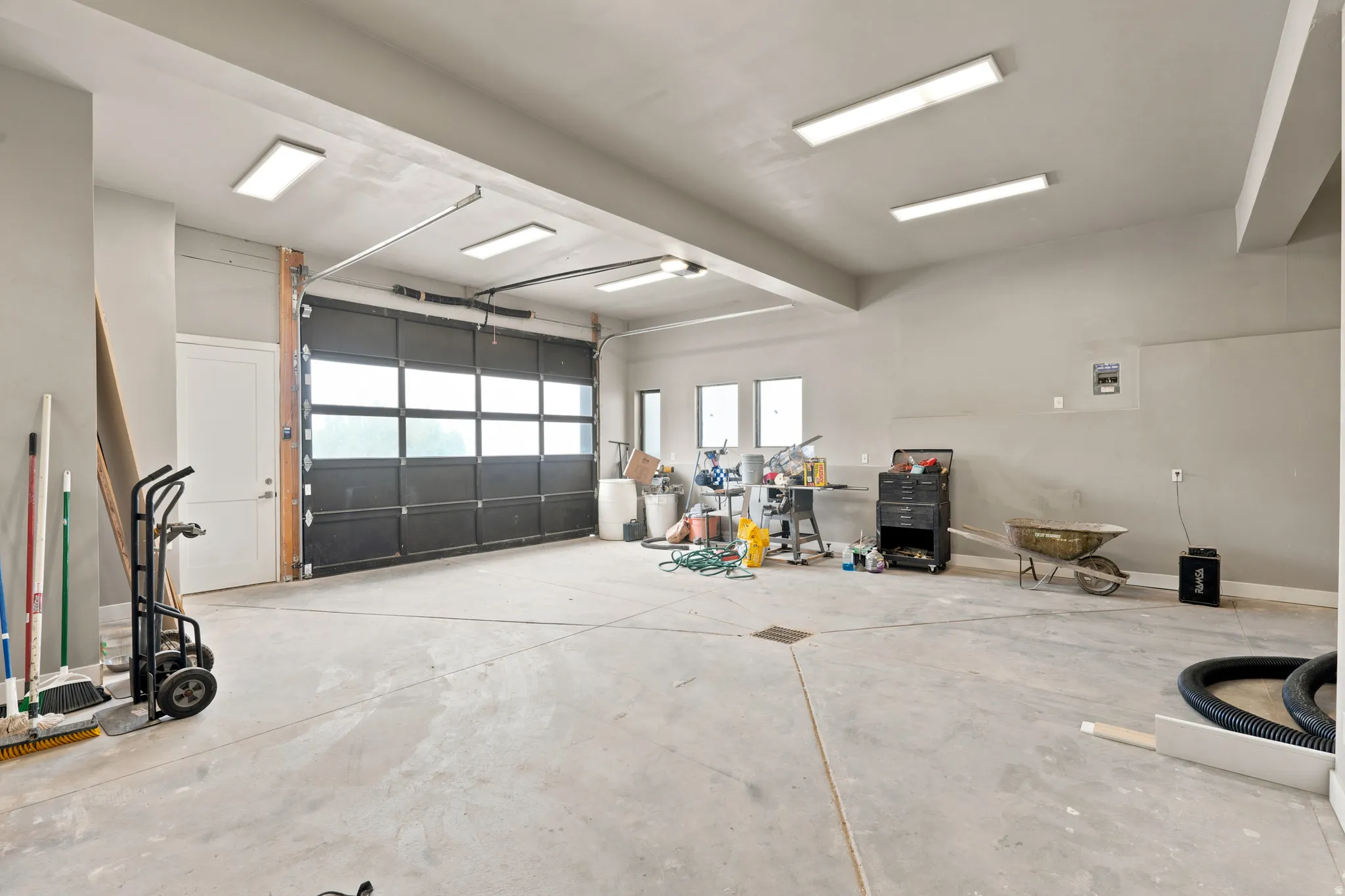 Spacious garage featuring a large glass-paneled overhead door, concrete flooring, multiple ceiling-mounted light fixtures, and a white side entry door