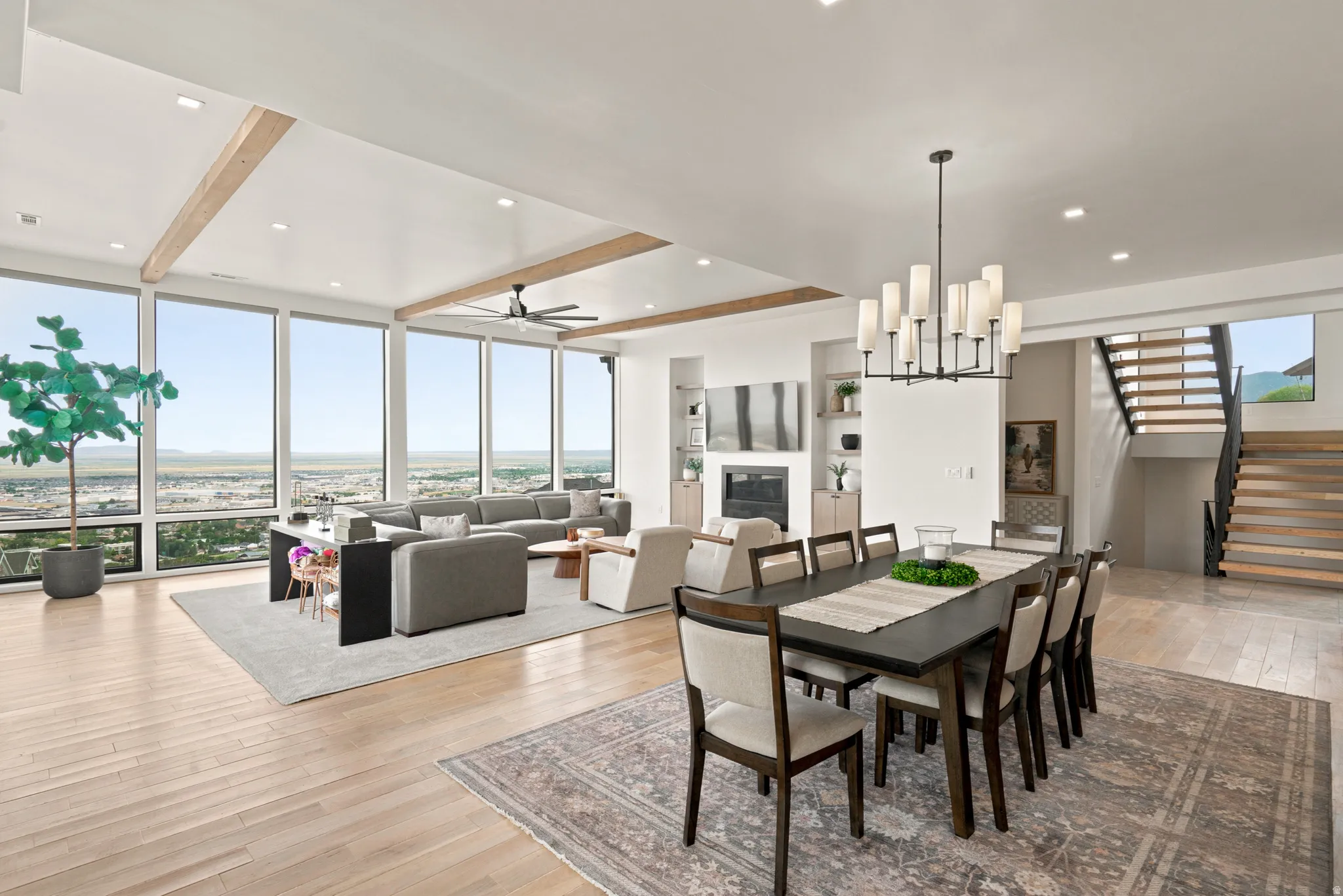 Expansive open-concept living area featuring light wood-finish flooring, a wall of floor-to-ceiling windows, and exposed ceiling beams