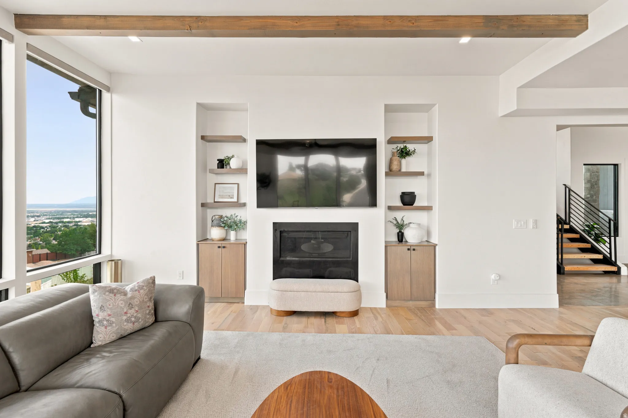 Spacious living area featuring a fireplace with built-in shelving and cabinetry, wood-finish flooring, exposed wood ceiling beams, and expansive windows