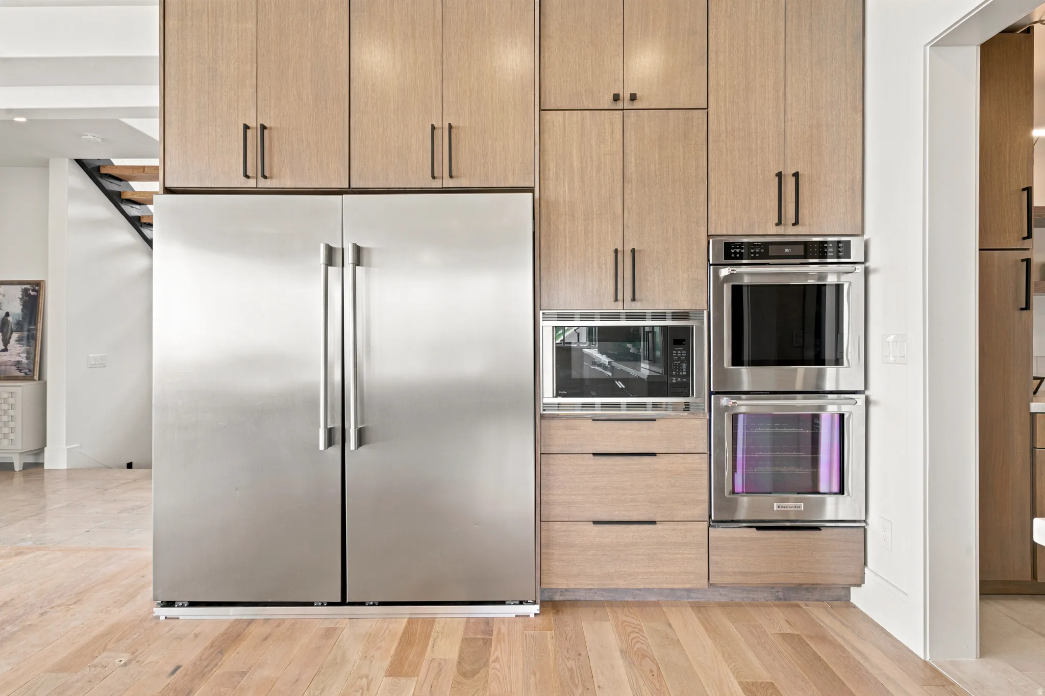 Gourmet kitchen appliance wall featuring a stainless steel side-by-side refrigerator, built-in microwave, and double wall ovens, complemented by wood-finish cabinetry and drawers