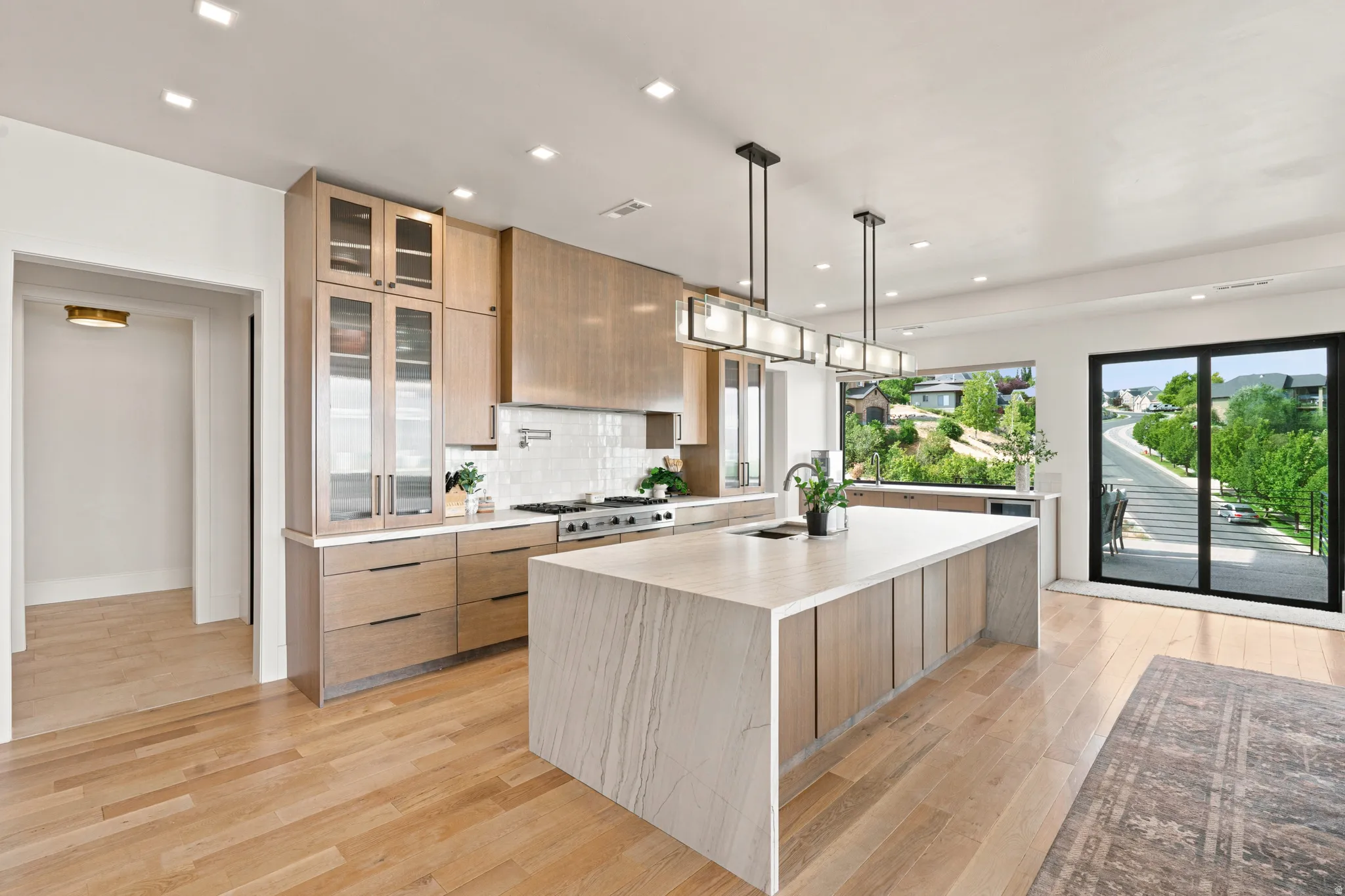 Modern kitchen featuring a large waterfall island, light wood-finish cabinetry, stainless steel appliances, and a tile backsplash