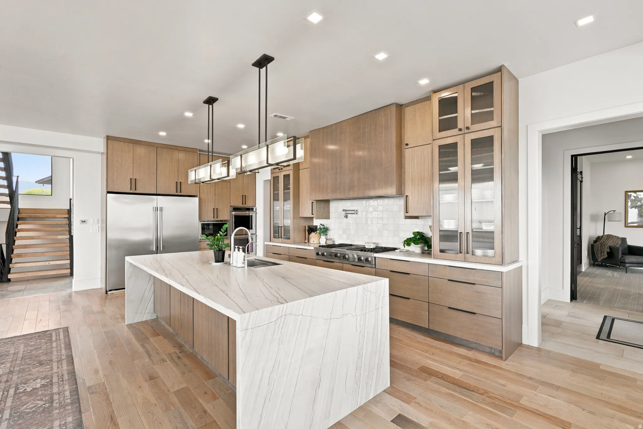 Modern kitchen featuring a waterfall island with a white patterned countertop, light wood cabinetry, stainless steel appliances, and wood-finish flooring
