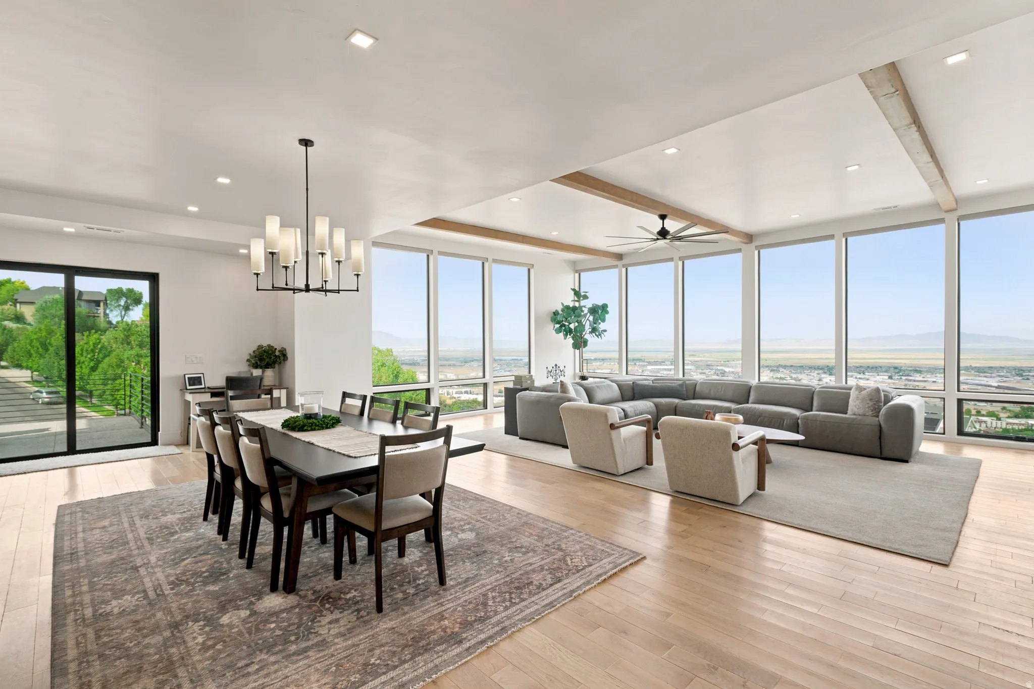 Expansive open-concept living area featuring a wall of floor-to-ceiling windows, wood-finish flooring, recessed lighting, exposed ceiling beams, and a modern chandelier