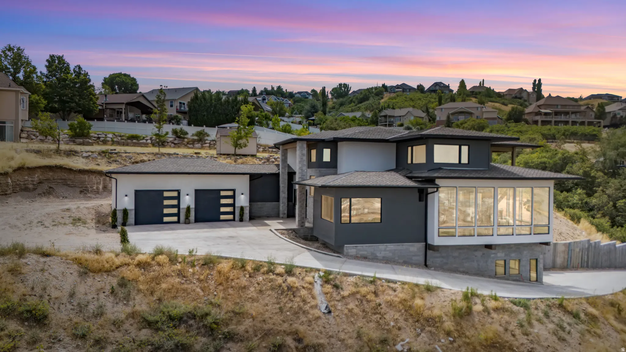 Modern architectural residence featuring multi-level design, a two-car garage with contemporary doors, extensive floor-to-ceiling windows, a stone-clad foundation, and a sloped concrete driveway