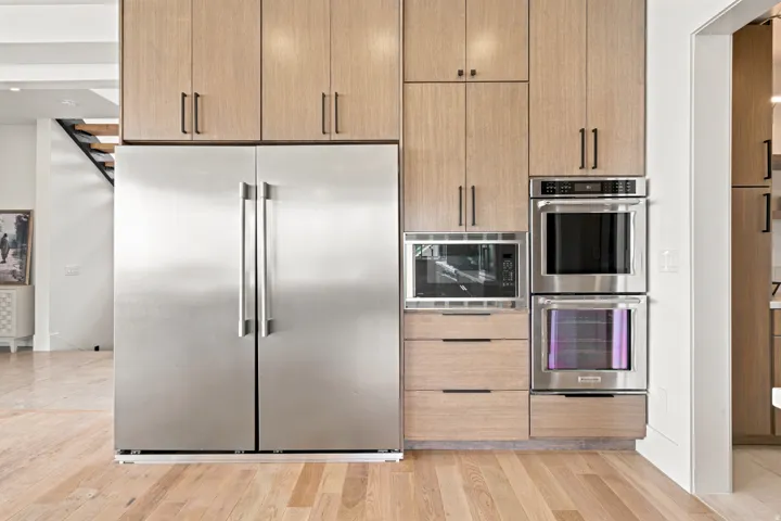 Gourmet kitchen appliance wall featuring a stainless steel side-by-side refrigerator, built-in microwave, and double wall ovens, complemented by wood-finish cabinetry and drawers