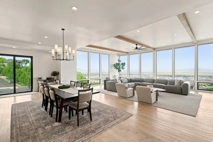 Expansive open-concept living area featuring a wall of floor-to-ceiling windows, wood-finish flooring, recessed lighting, exposed ceiling beams, and a modern chandelier