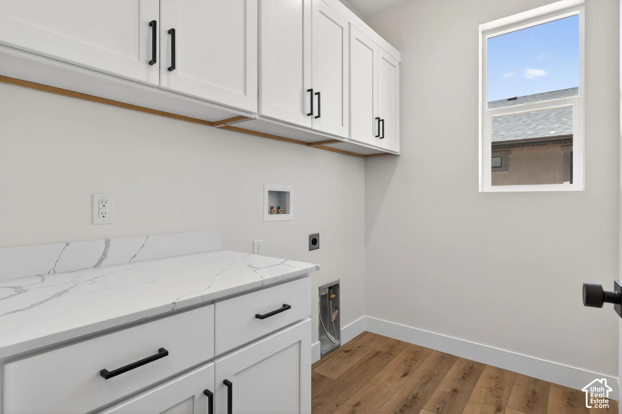 Laundry area featuring white shaker-style cabinetry with matte black hardware, a light-colored countertop with veining, and wood-finish flooring