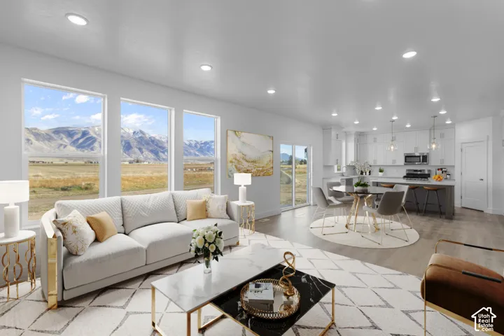 Bright, open-concept living area showcasing expansive windows framing mountain and valley scenery, light wood-finish flooring throughout, a neutral upholstered sofa with coordinating accent pillows, a contemporary glass-top coffee table, recessed ceiling