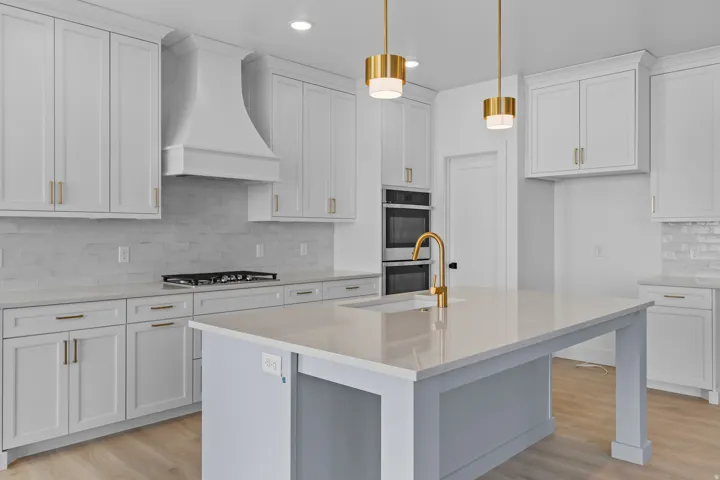 Sophisticated kitchen featuring white shaker cabinetry with brass hardware, a large waterfall-edge island with a gray base and white quartz countertop, stainless steel gas cooktop, dual wall-mounted ovens, a professional-style range hood, pendant lighting