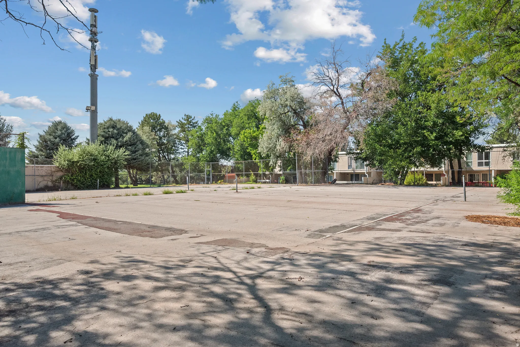 Asphalt surface court with chain-link fencing, surrounded by mature trees and residential structures in the background