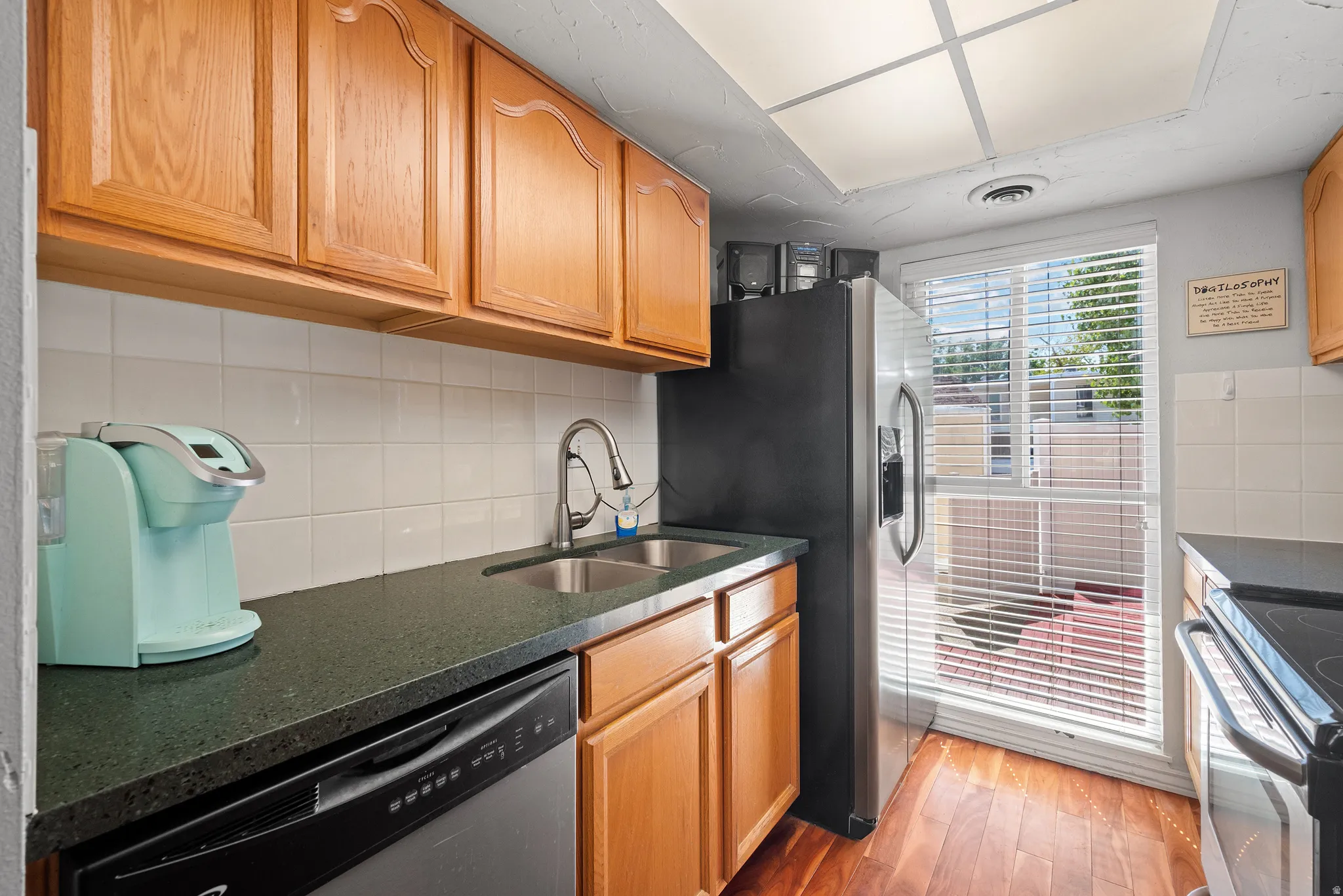 Kitchen featuring wood-finish flooring, wood cabinetry, dark countertops, a tile backsplash, and a double basin stainless steel sink