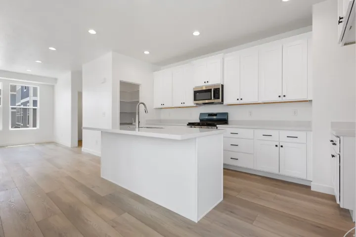 Open-concept kitchen featuring an island with an undermount sink and gooseneck faucet, white shaker-style cabinetry, light-toned countertops, and stainless steel appliances