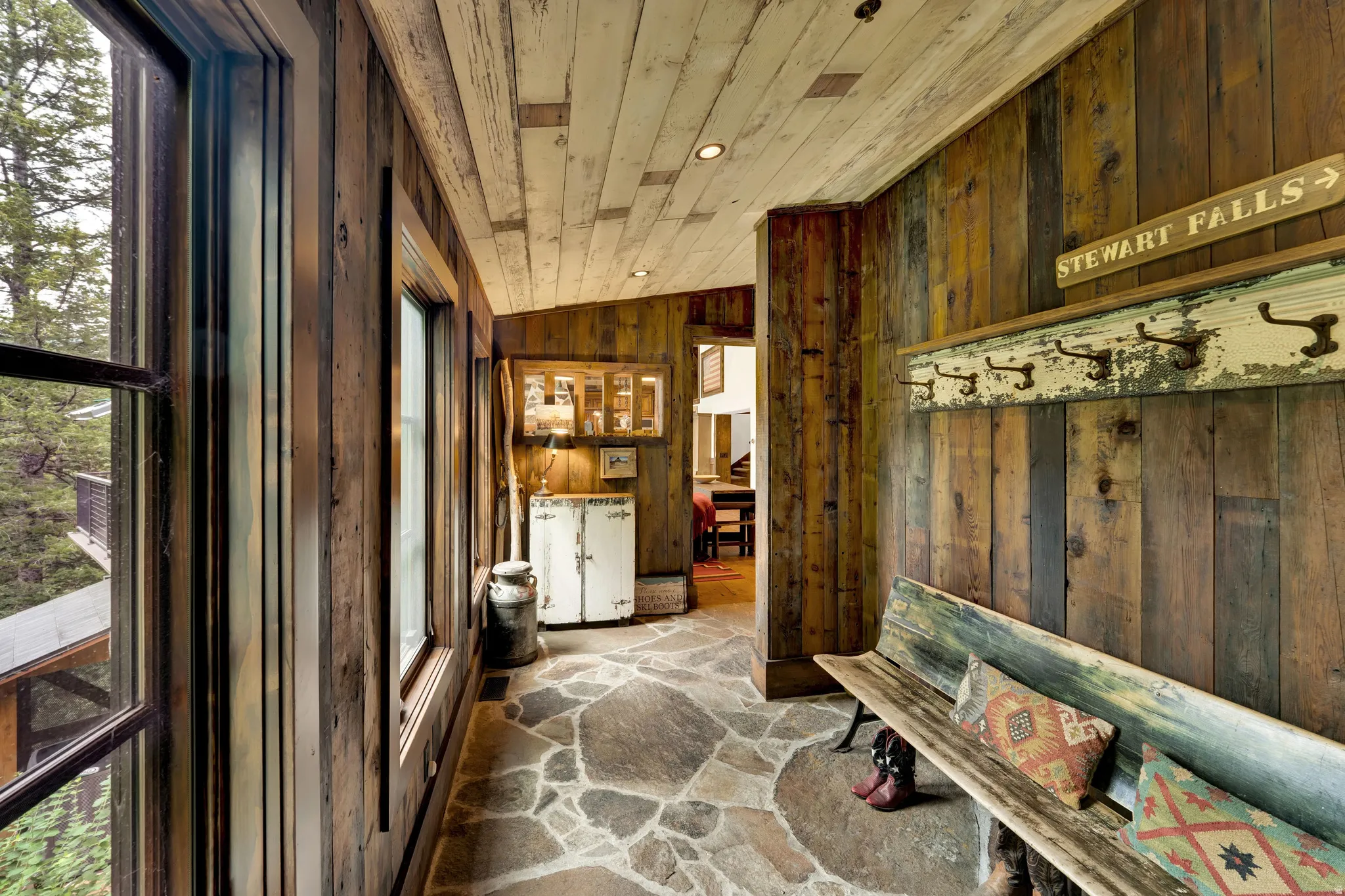 Rustic mudroom entry featuring reclaimed wood paneling throughout walls and ceiling, natural stone flooring with geometric patterning, large multi-pane windows with dark frames, vintage signage and architectural salvage displays, and direct access to adja
