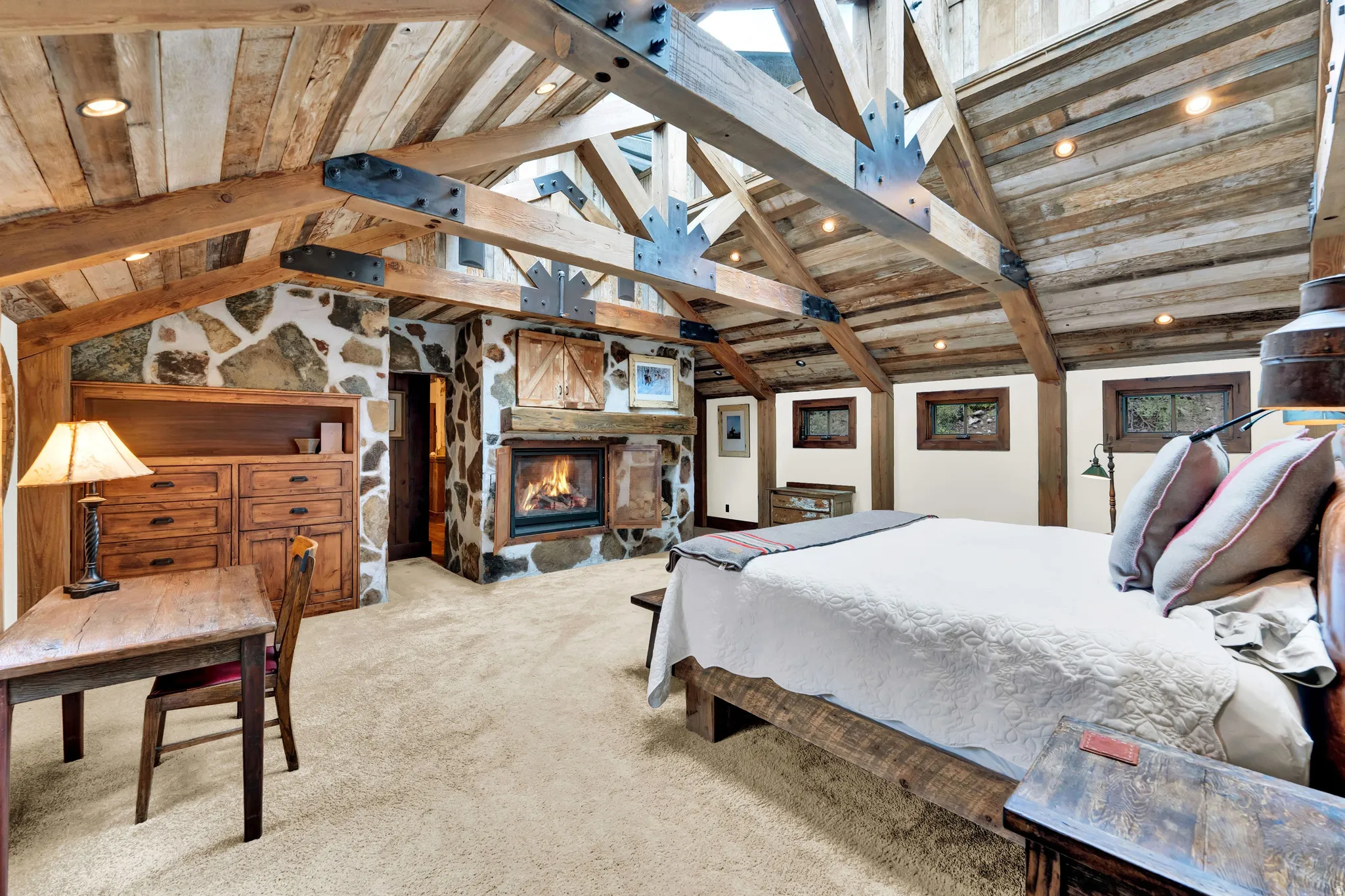 Spacious primary bedroom showcasing soaring vaulted ceilings with exposed wooden beams, skylights flooding the space with natural light, and a wood-burning fireplace