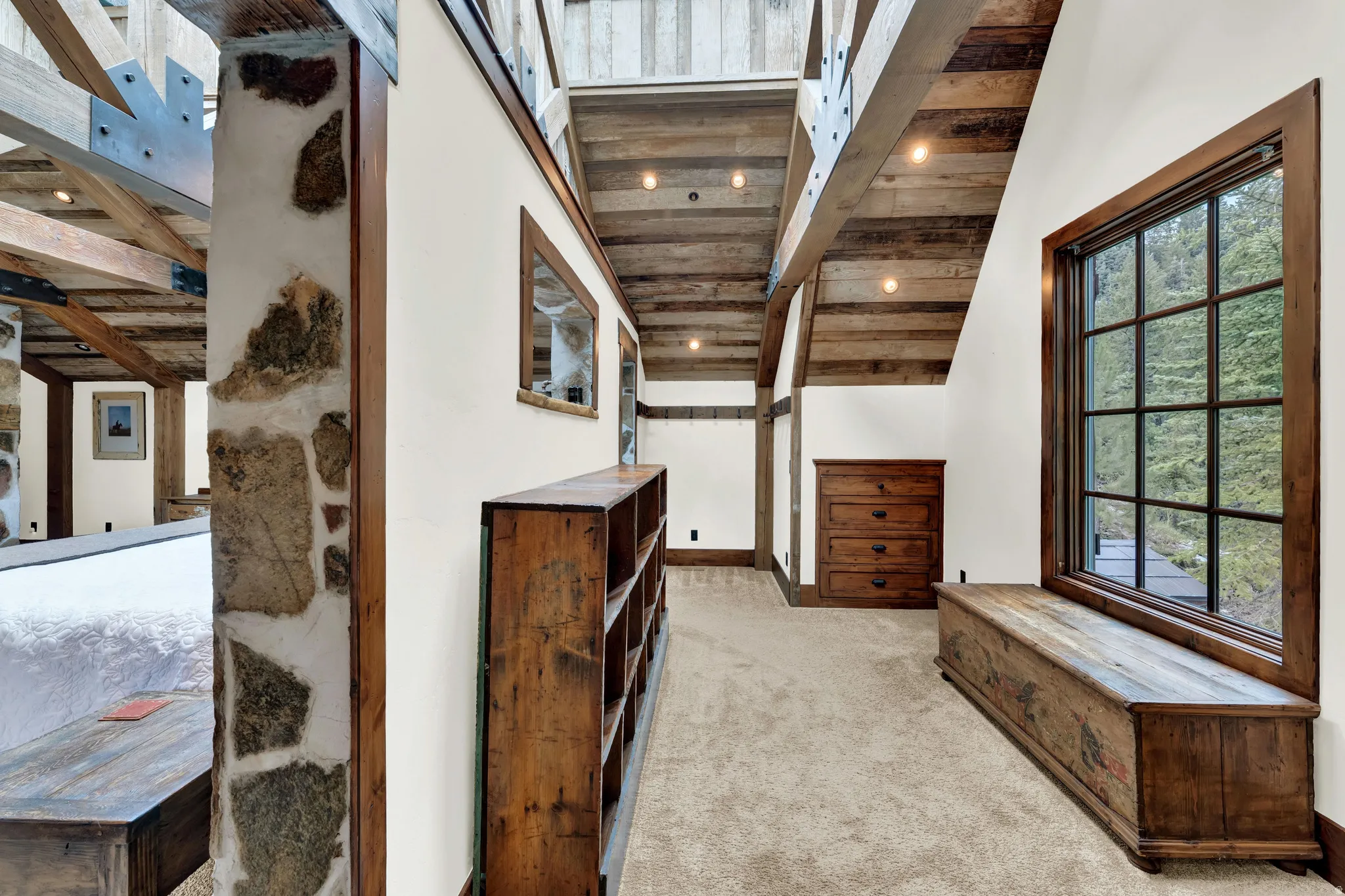 Hallway featuring exposed wood beam ceiling details, natural light from large multi-pane windows, wood-finish cabinetry, stone accent walls, and neutral carpeting throughout