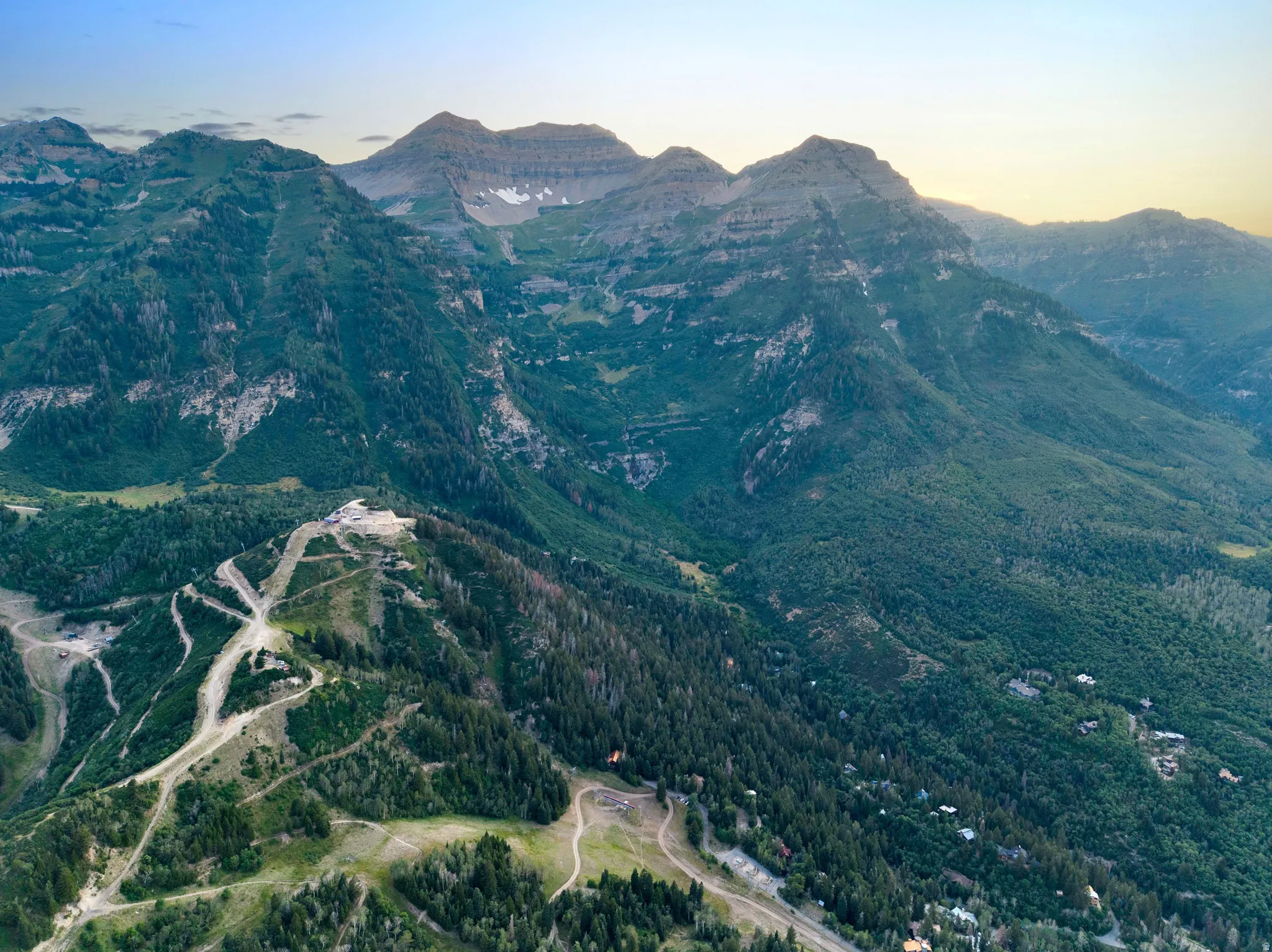 Scenic mountain valley setting featuring dense evergreen forest coverage, winding road infrastructure through the landscape, and layered alpine peaks in the distance