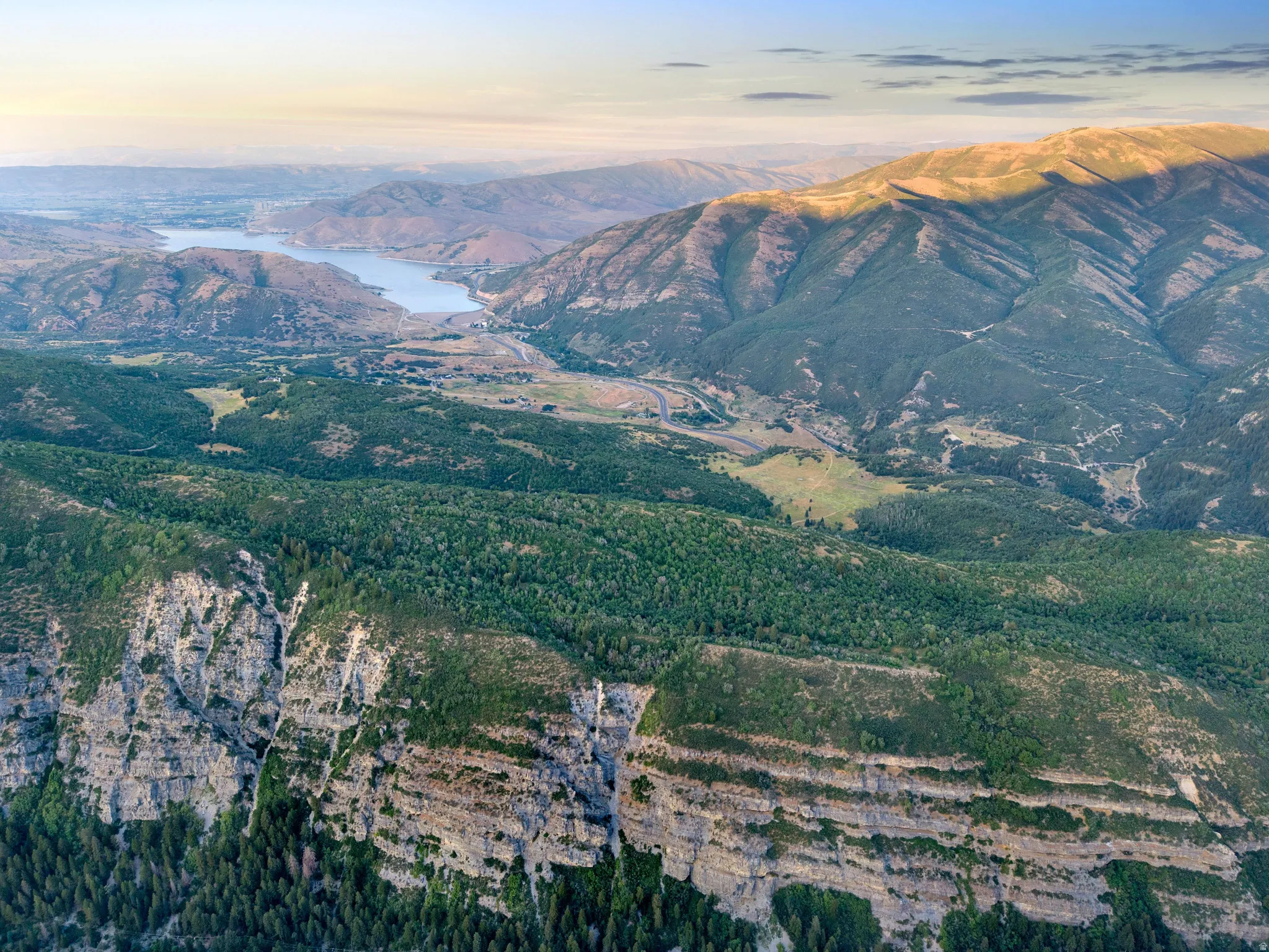 Aerial view showcasing expansive forested valleys with dramatic layered ridgelines, a pristine alpine lake, and rolling terrain dotted with coniferous trees