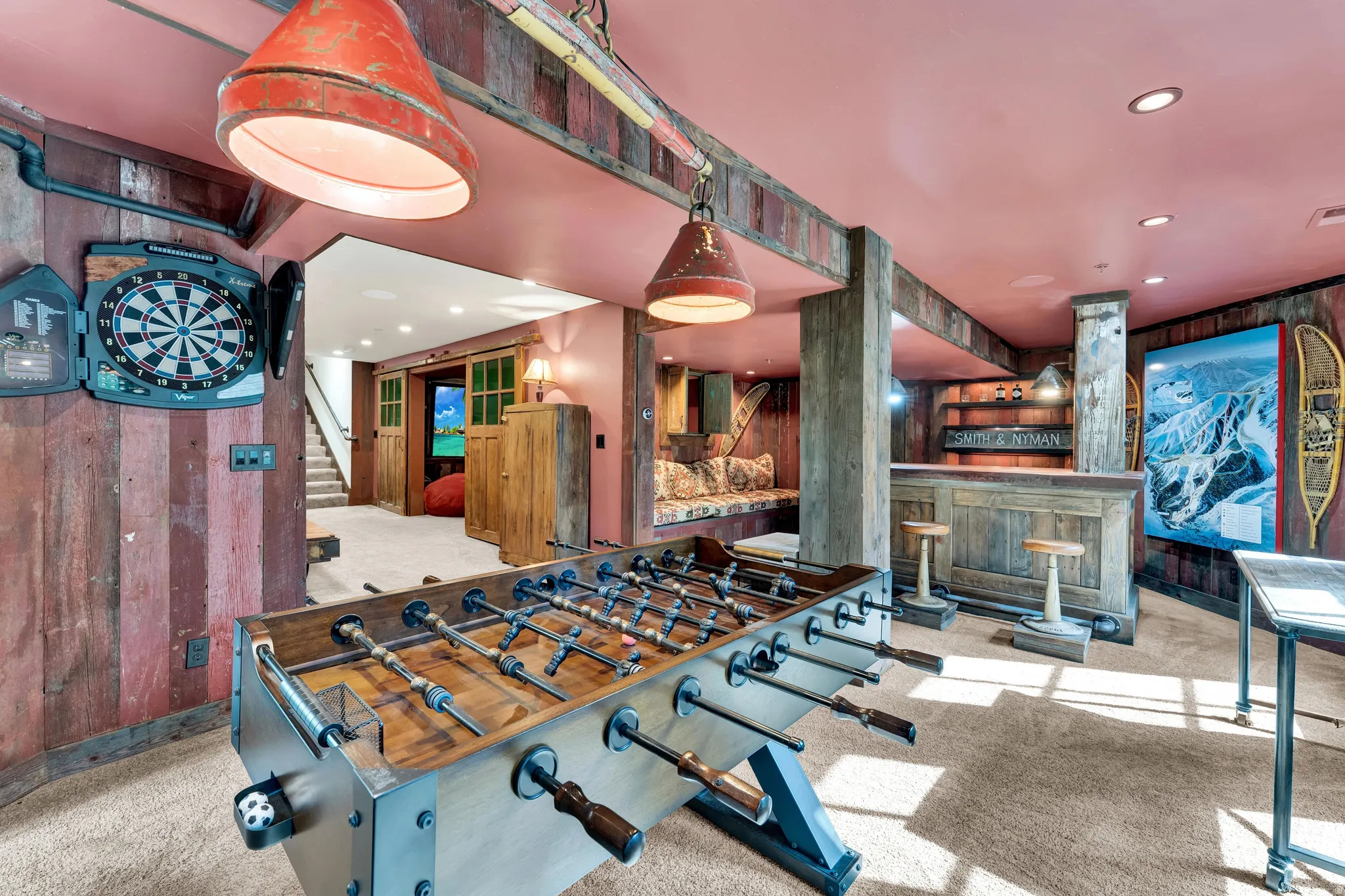 Reclaimed wood paneling and exposed beams accent this entertainment space featuring recessed lighting, a built-in wet bar with shelving, a window seat, and a foosball table