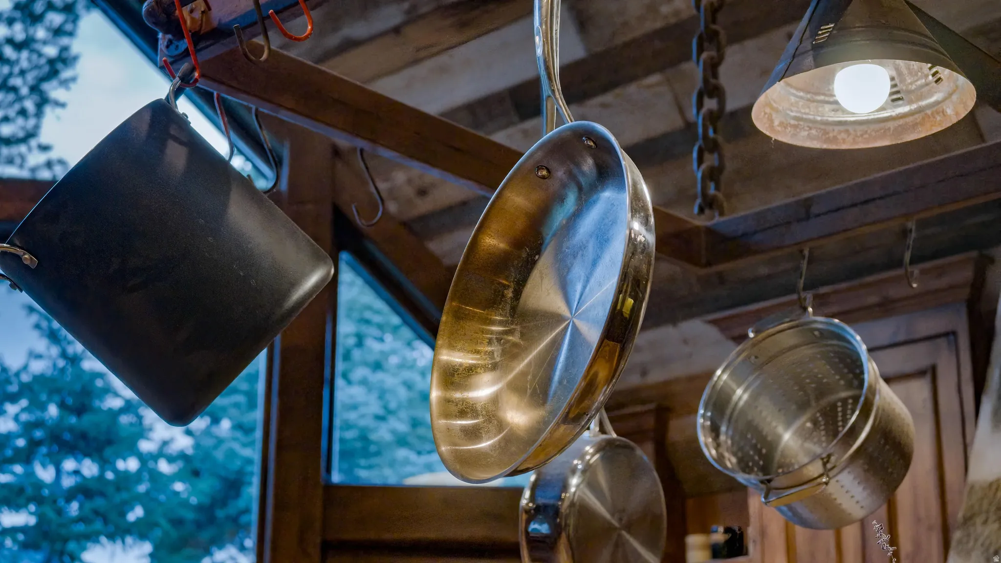 Rustic kitchen featuring exposed wooden ceiling beams, stainless steel cookware hanging from chains, and pendant lighting with warm brass-toned shades