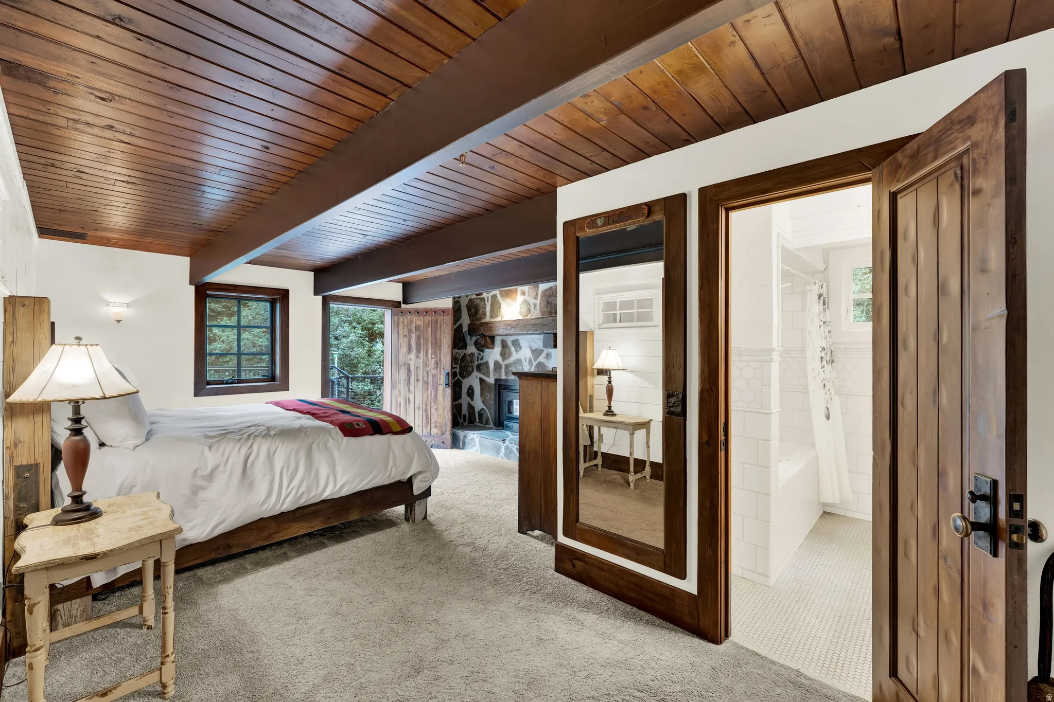 Carpeted sleeping quarters featuring a wood-plank ceiling with exposed beams, a built-in stone fireplace, wood-framed windows, and a private entryway to a bathroom with white tile flooring and a porcelain tub