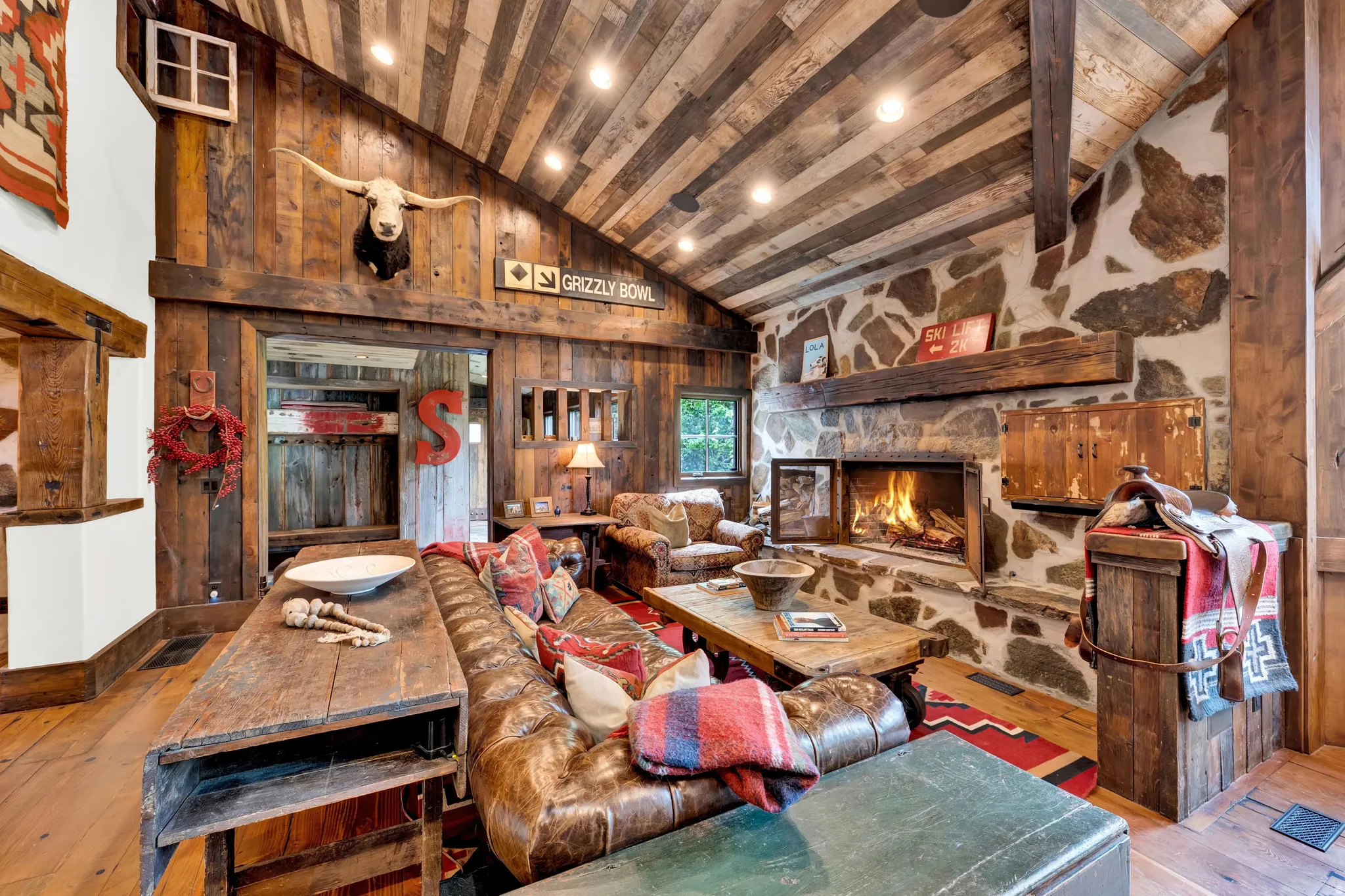 Rustic timber-beam great room featuring exposed wood-finish vaulted ceilings, a wood-burning fireplace with stone surround, and wood-finish flooring throughout