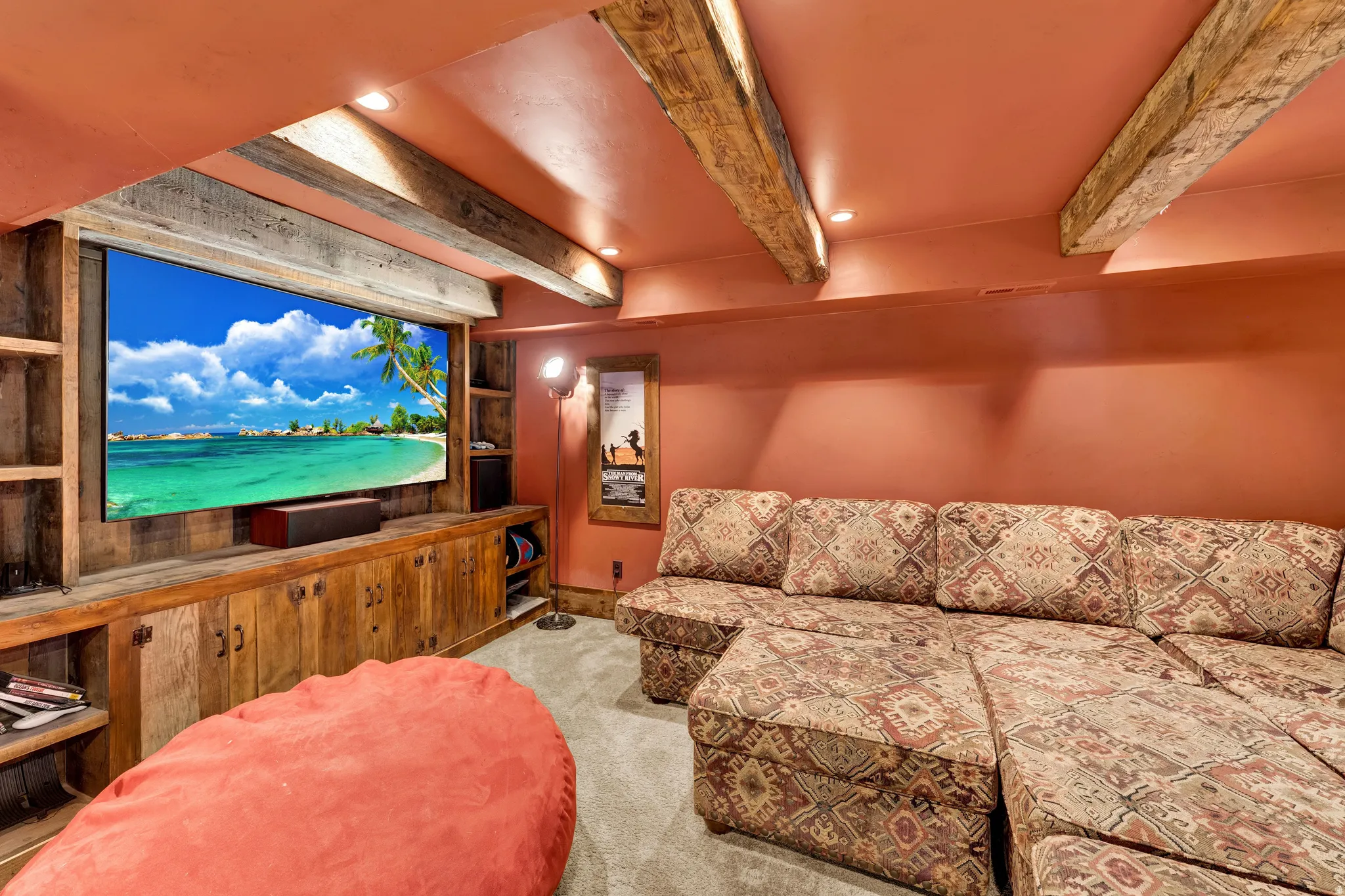 Dedicated media room featuring exposed wood beam ceiling with recessed lighting, custom wood cabinetry with storage, large-format display screen, plush seating with patterned upholstery, and concrete flooring throughout