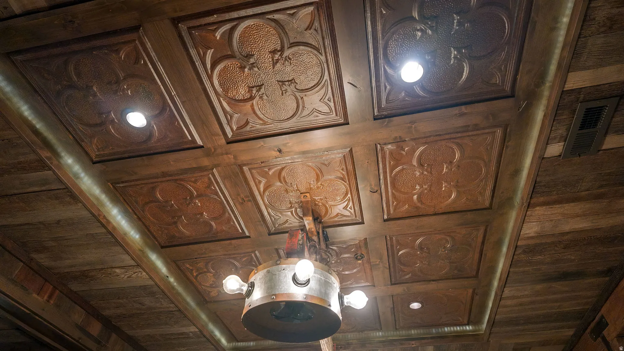 Coffered ceiling with decorative carved wood panels and integrated pendant lighting fixture featuring multiple light sources and brushed metal accents