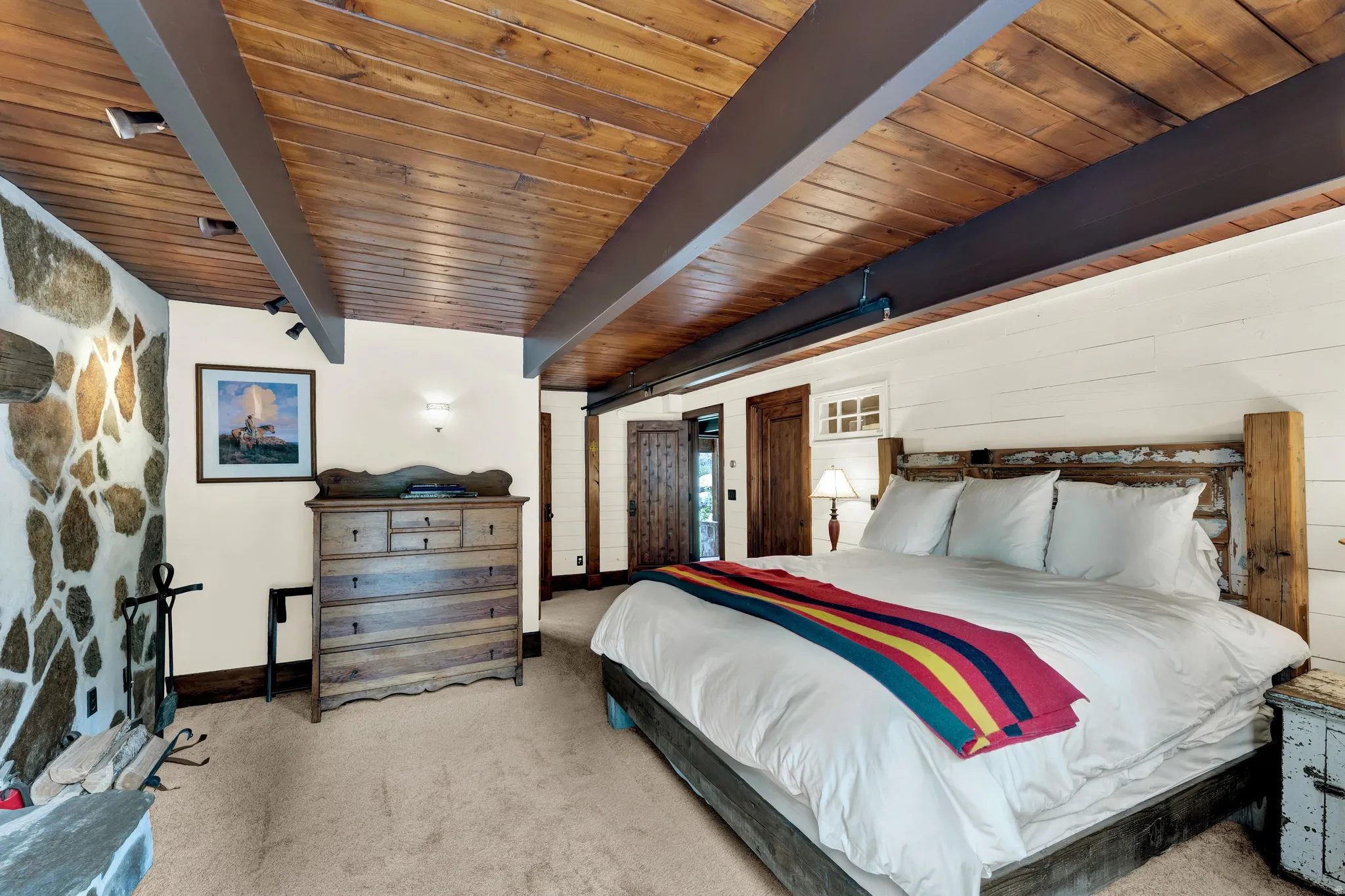 Spacious primary bedroom featuring exposed wood-beam ceiling with recessed lighting, stone accent wall, hardwood flooring, and a wooden bed frame with white bedding