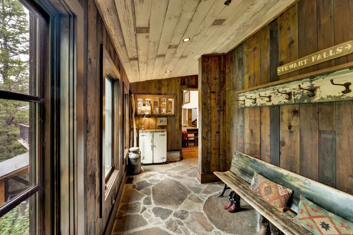 Rustic mudroom entry featuring reclaimed wood paneling throughout walls and ceiling, natural stone flooring with geometric patterning, large multi-pane windows with dark frames, vintage signage and architectural salvage displays, and direct access to adja