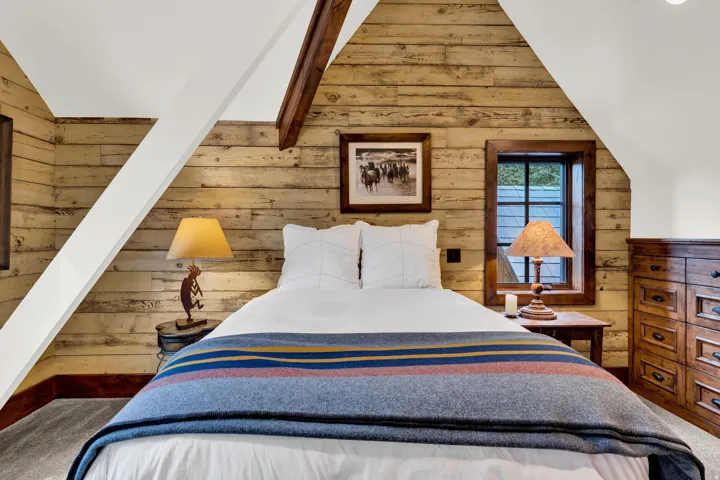 Charming bedroom retreat showcasing rustic wood plank walls, vaulted ceilings with exposed beam detailing, and abundant natural light from dual windows