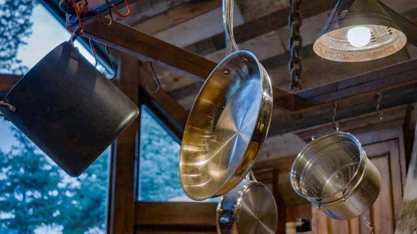 Rustic kitchen featuring exposed wooden ceiling beams, stainless steel cookware hanging from chains, and pendant lighting with warm brass-toned shades