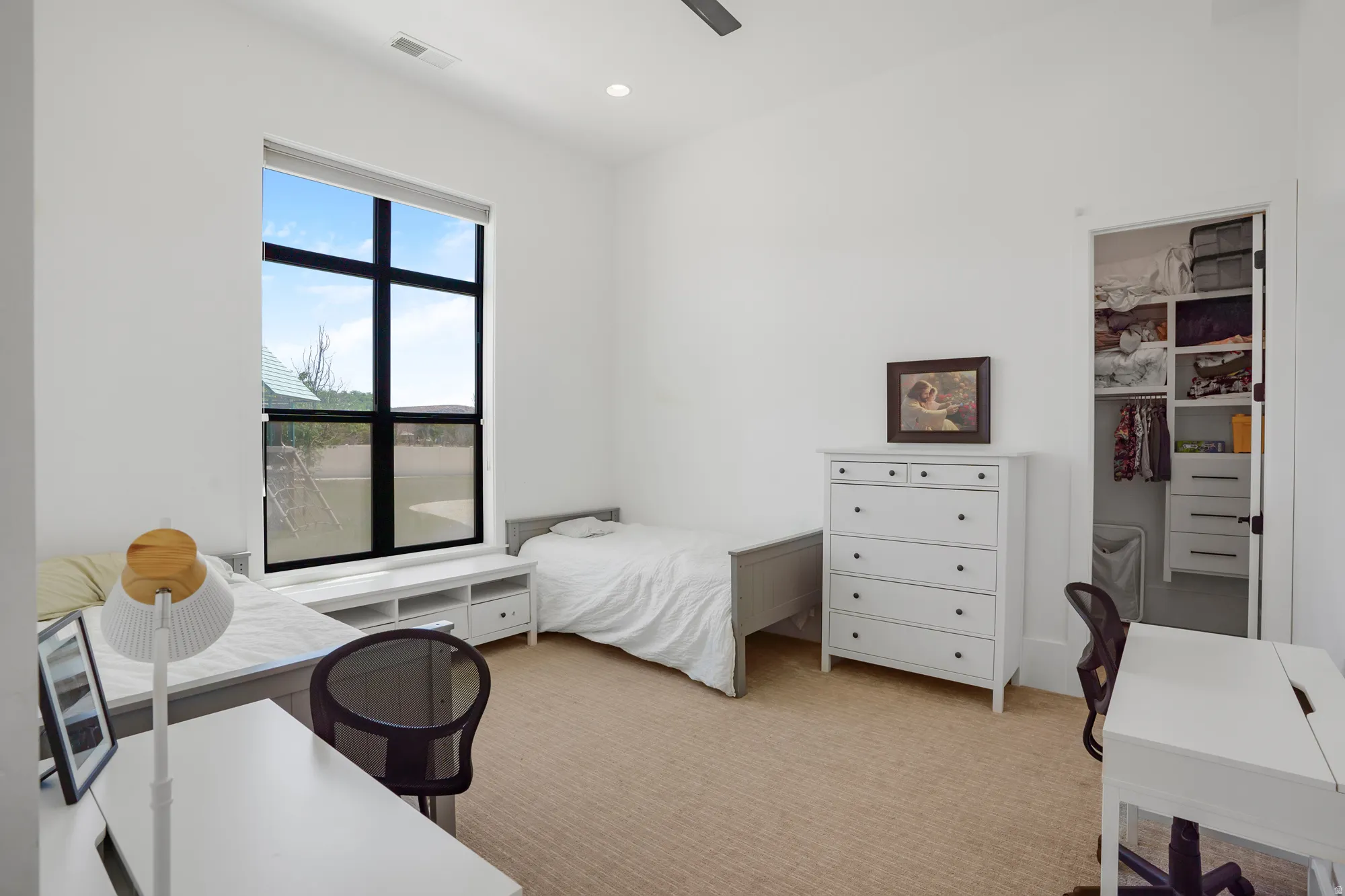 Bright bedroom suite featuring large black-framed windows with natural light, white painted wood dresser with multiple drawers, built-in closet with shelving and organizational systems, wood-finish flooring, and ample space for bedroom furnishings and a w