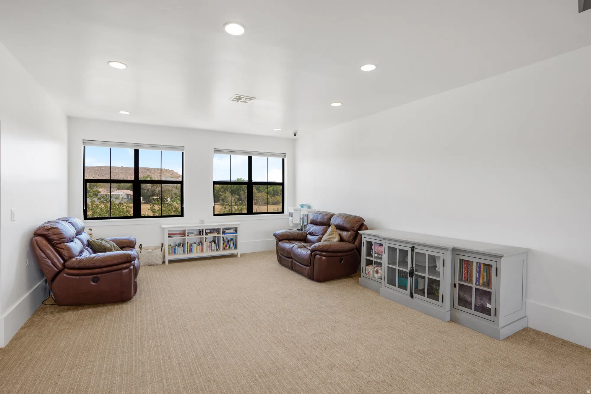 Bright and spacious recreation room featuring multiple recessed ceiling lights, large multi-pane windows with scenic outlooks, wood-finish flooring throughout, and built-in shelving with glass-front cabinetry for display and storage