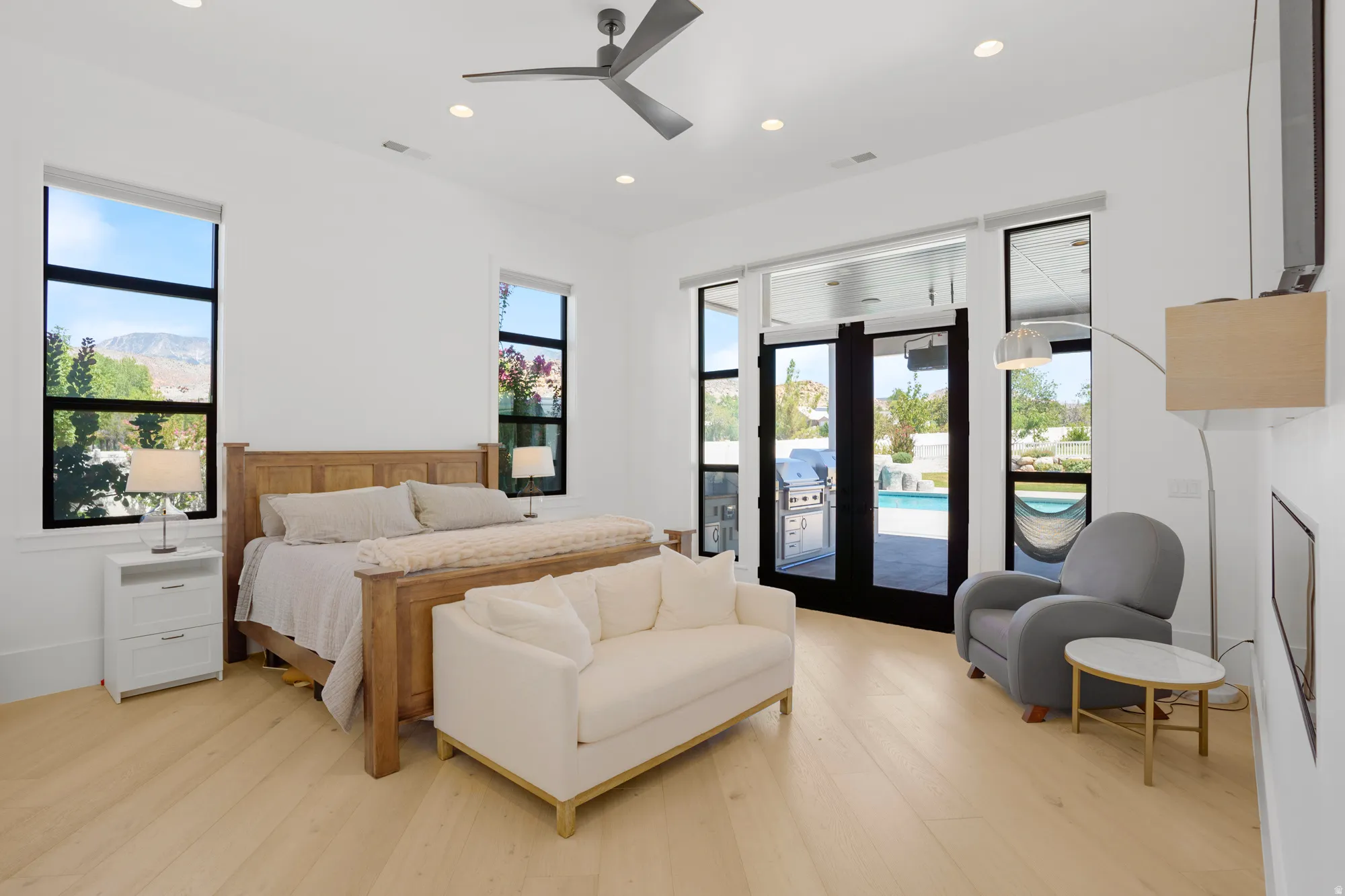 Spacious primary bedroom featuring a wood-frame bed, wood-finish flooring throughout, abundant natural light from multiple windows, and glass sliding doors opening to outdoor space