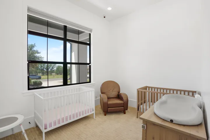 Bright nursery featuring abundant natural light from a large black-framed window, light wood-finish flooring, and ample space for multiple cribs and furnishings