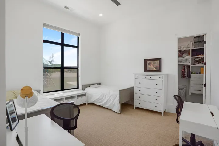 Bright bedroom suite featuring large black-framed windows with natural light, white painted wood dresser with multiple drawers, built-in closet with shelving and organizational systems, wood-finish flooring, and ample space for bedroom furnishings and a w